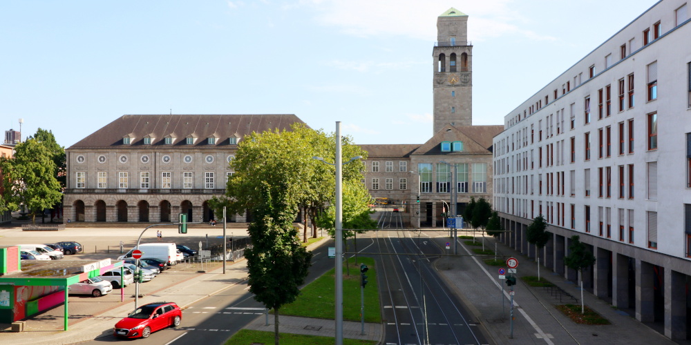 Ein langgezogenes historisches Gebäude mit einem Platz davor, das rechts über die Straße geführt wird, dahinter ein Turm